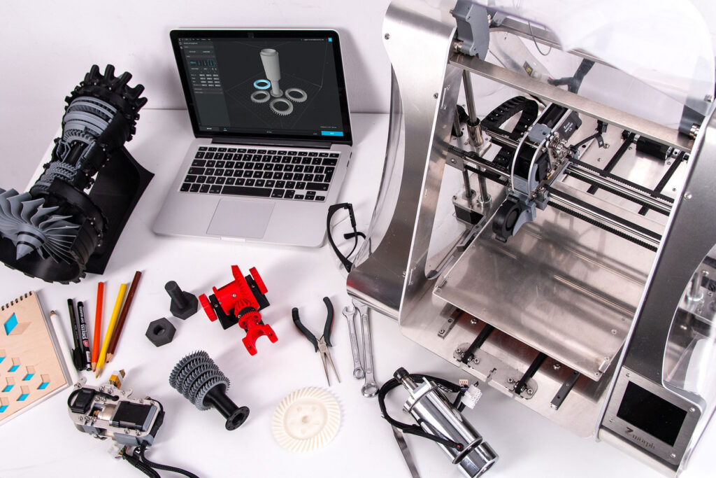 3D Printer Setup A picture of a white table containing a small laptop, a 3D-printer, several 3D-printed objects in glack, grey, and several tools, pencils, and writing pad.